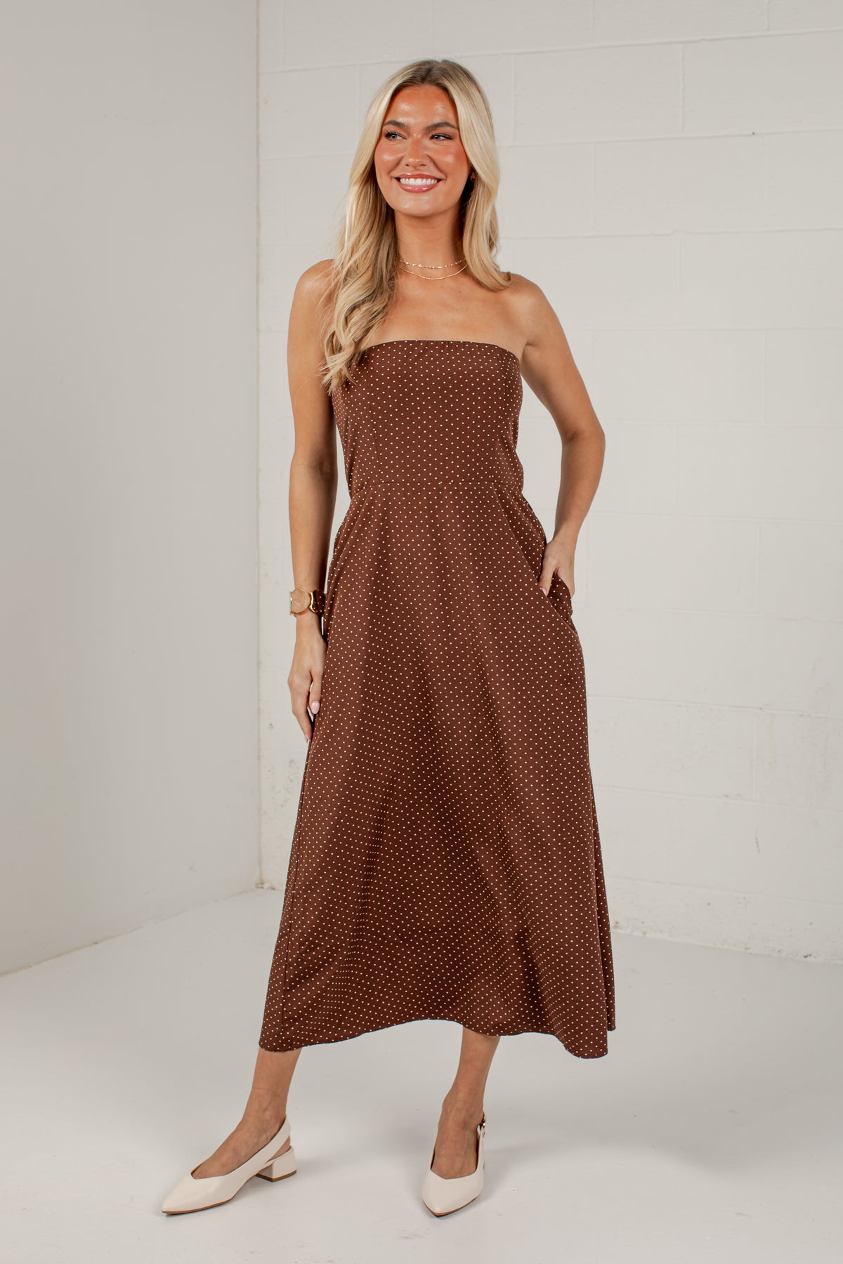 A woman modeling a brown strapless midi dress with a small white polka dot pattern, standing against a white brick wall.