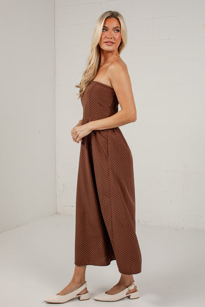 A woman modeling a brown strapless jumpsuit with a small white polka dot pattern, paired with white Mary Jane shoes, standing in a white studio corner.