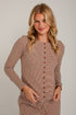 A woman modeling a long-sleeve knit top with thin brown and beige horizontal stripes, featuring a button-down front and a drawstring waist, set against a light gray brick wall.