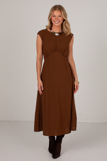 A woman modeling a sleeveless brown midi dress with a pleated texture, standing against a plain light gray studio background.