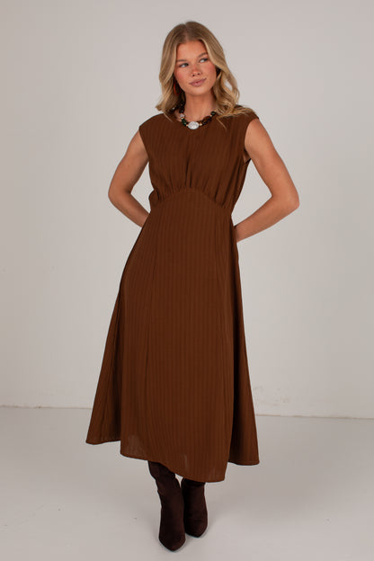 A woman modeling a sleeveless brown midi dress with a pleated texture, standing against a plain light gray studio background.