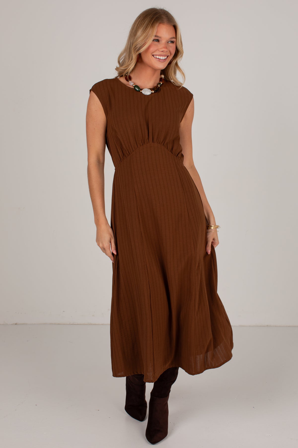 A woman modeling a sleeveless brown midi dress with a gathered waist and pleated texture, paired with dark brown ankle boots against a plain light gray studio background.