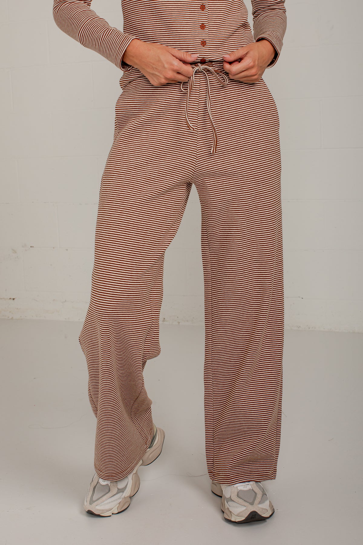 A person wearing loose, wide-leg knit pants with thin brown and beige horizontal stripes, paired with a matching long-sleeve top and white sneakers, standing against a plain light gray wall.