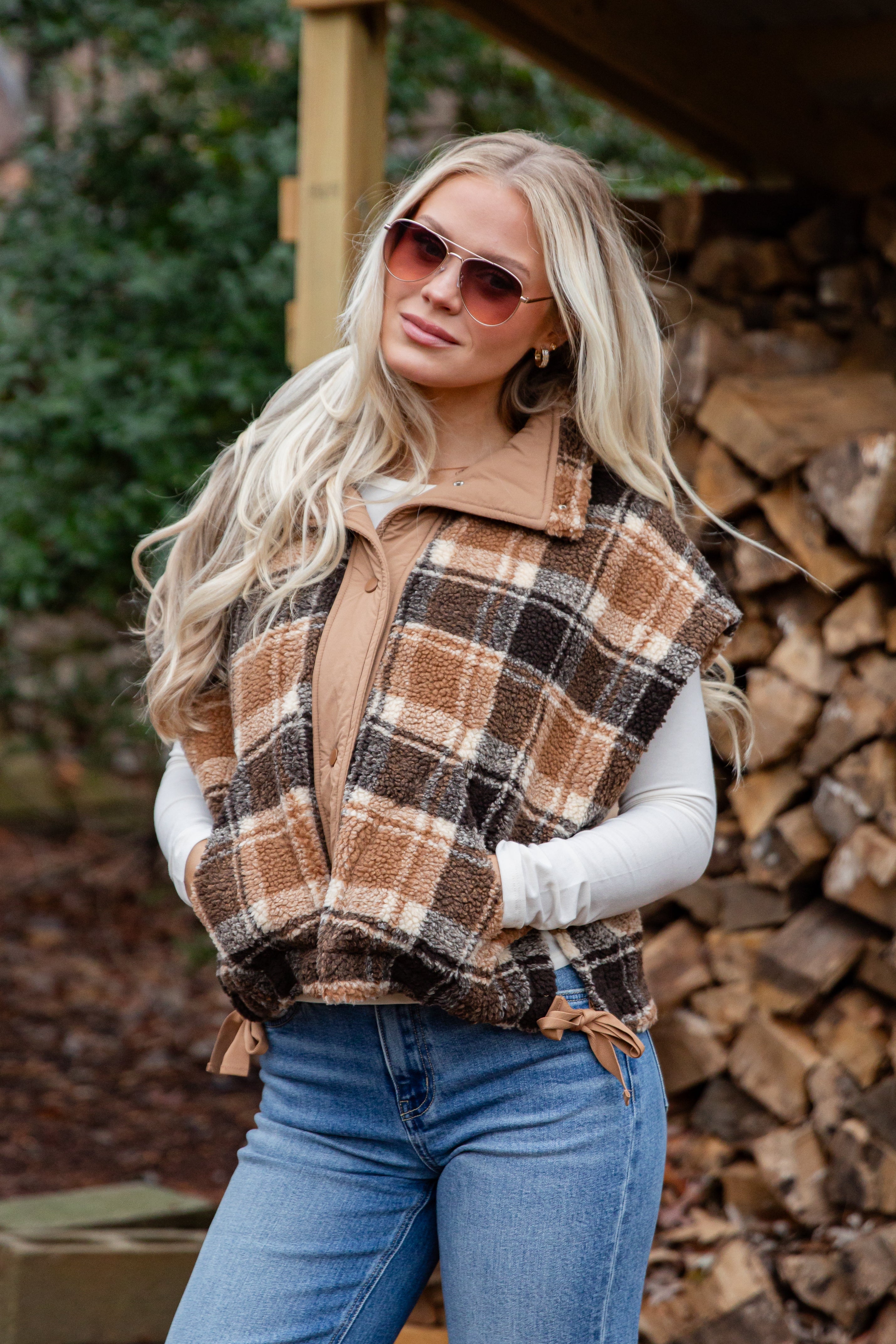 Woman wearing a plaid vest and sunglasses outdoors with stacked firewood in the background