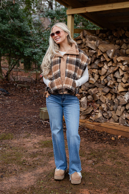 Woman wearing a plaid vest and blue jeans standing in front of stacked firewood.