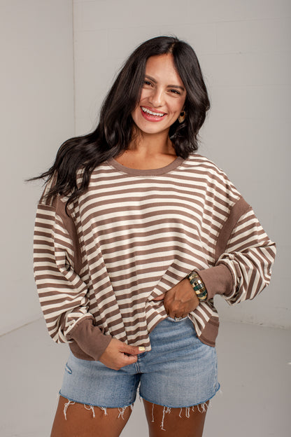 A woman wearing a brown and beige striped long-sleeve top with solid brown contrast panels on the shoulders, cuffs, and hem, paired with denim shorts, against a light gray studio background.