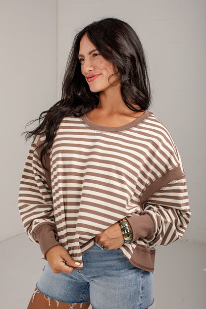A woman wearing a brown and beige horizontally striped long-sleeve top with solid brown ribbed trim, paired with denim shorts, standing against a plain light gray wall.