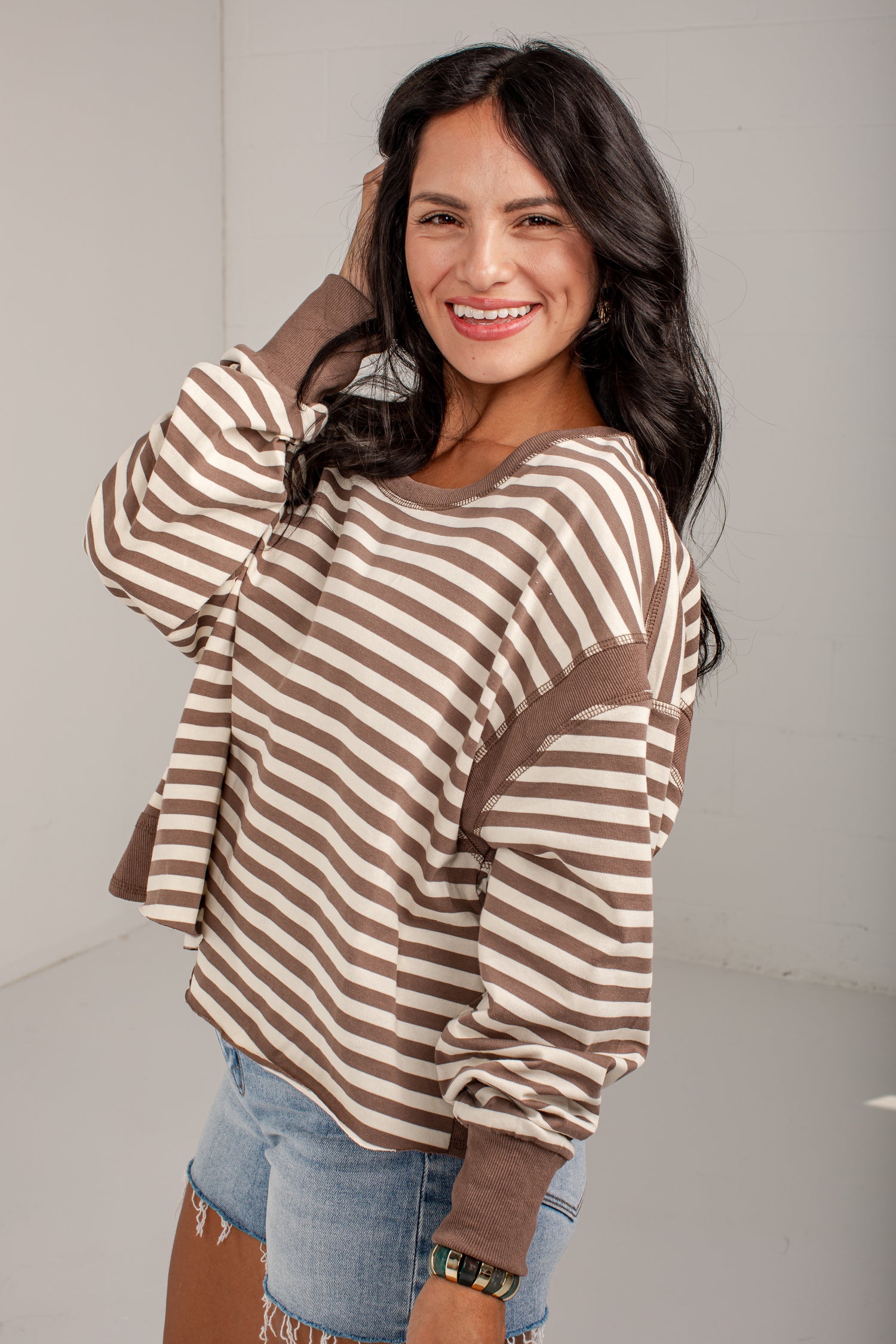 A woman modeling a brown and beige horizontally striped long-sleeve top with solid brown ribbed cuffs and hem, paired with light blue denim jeans.