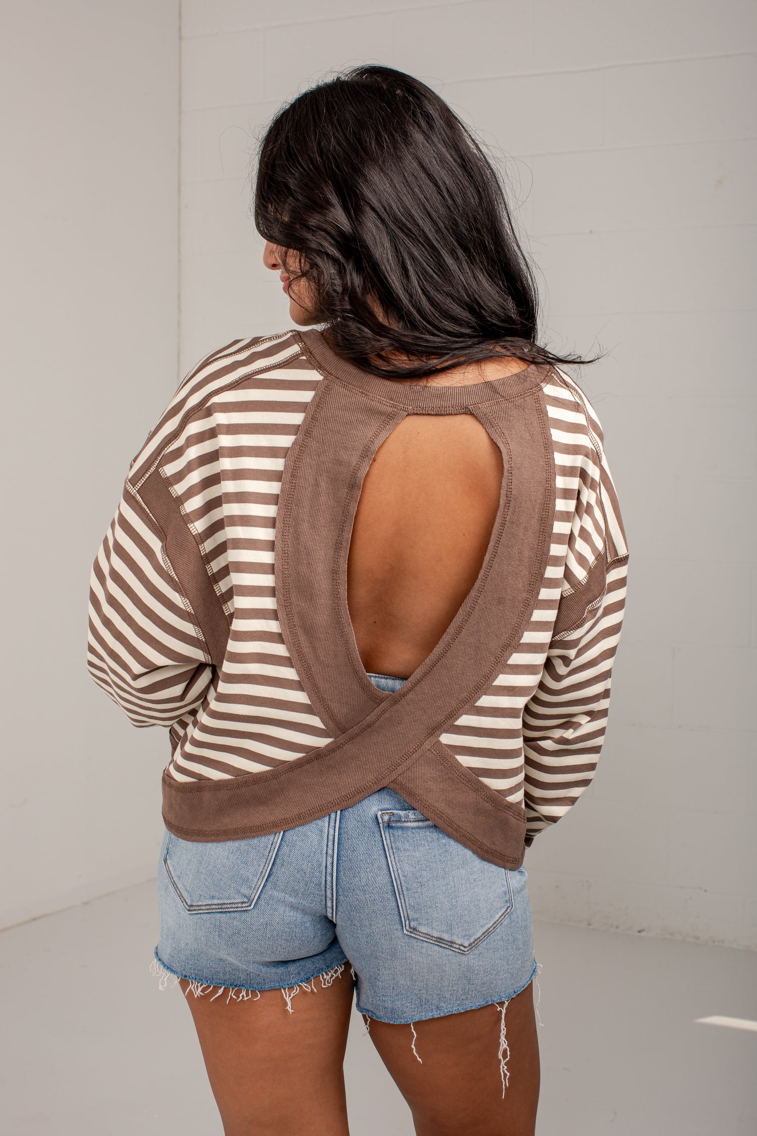 A woman shown from the back wearing a brown and beige striped long-sleeve top with a large crossover open back, paired with light blue denim shorts.