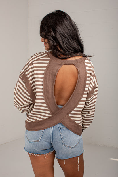 A woman shown from the back wearing a brown and beige striped long-sleeve top with a large crossover open back, paired with light blue denim shorts.