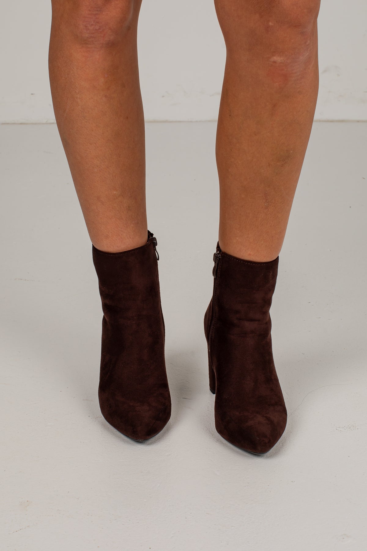A pair of dark brown suede ankle boots with block heels and side zippers, shown on a model&