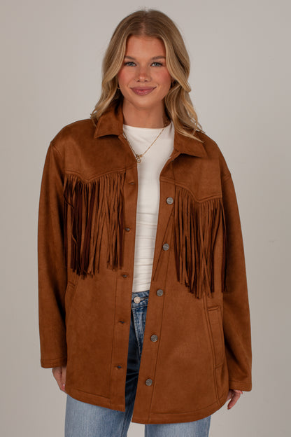 A woman modeling a brown faux-suede fringe jacket over a white top and blue jeans, photographed against a plain light gray studio background.
