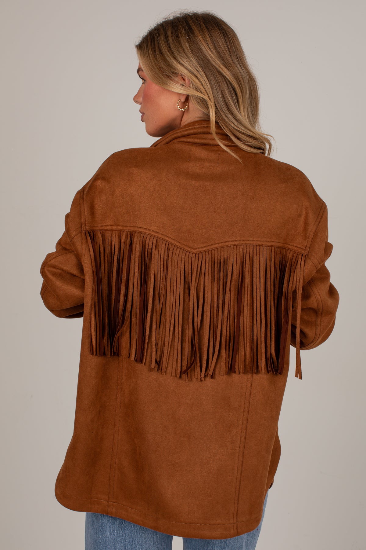 A woman shown from the back wearing a brown suede-like fringe jacket over a dark top and blue jeans, photographed against a plain light gray studio background.