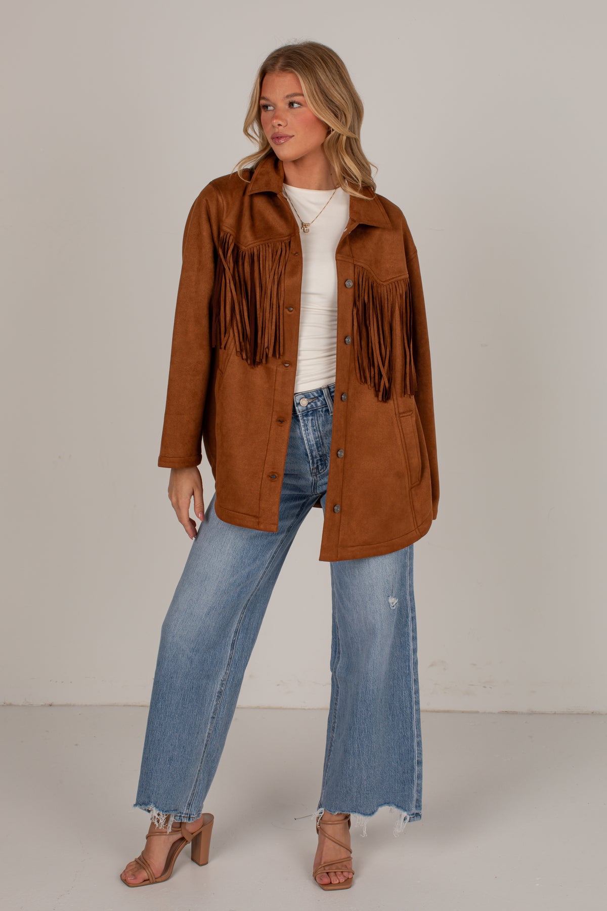 A woman modeling a brown suede fringe jacket over a white ribbed top and blue wide-leg jeans, standing against a plain light gray studio background.