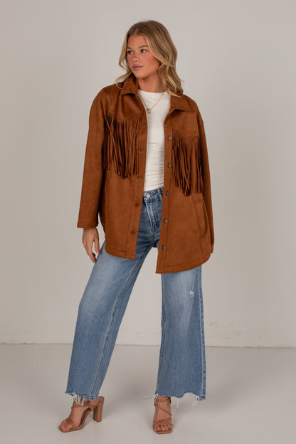 A woman modeling a brown suede fringe jacket over a white ribbed top and blue wide-leg jeans, standing against a plain light gray studio background.