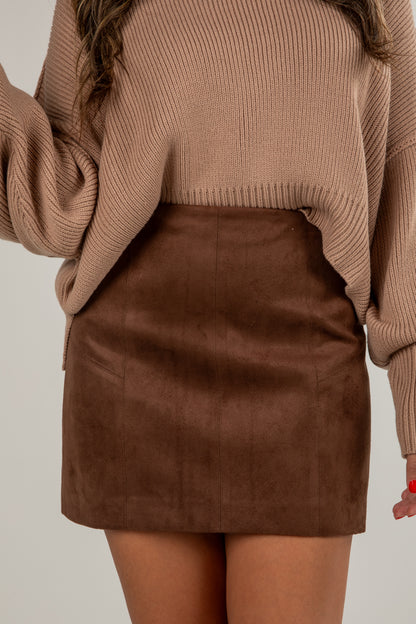 Person wearing a brown sweater and suede skirt on a plain background