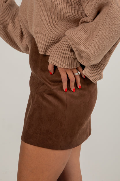 Person wearing a brown sweater and suede skirt with red nail polish on a plain background