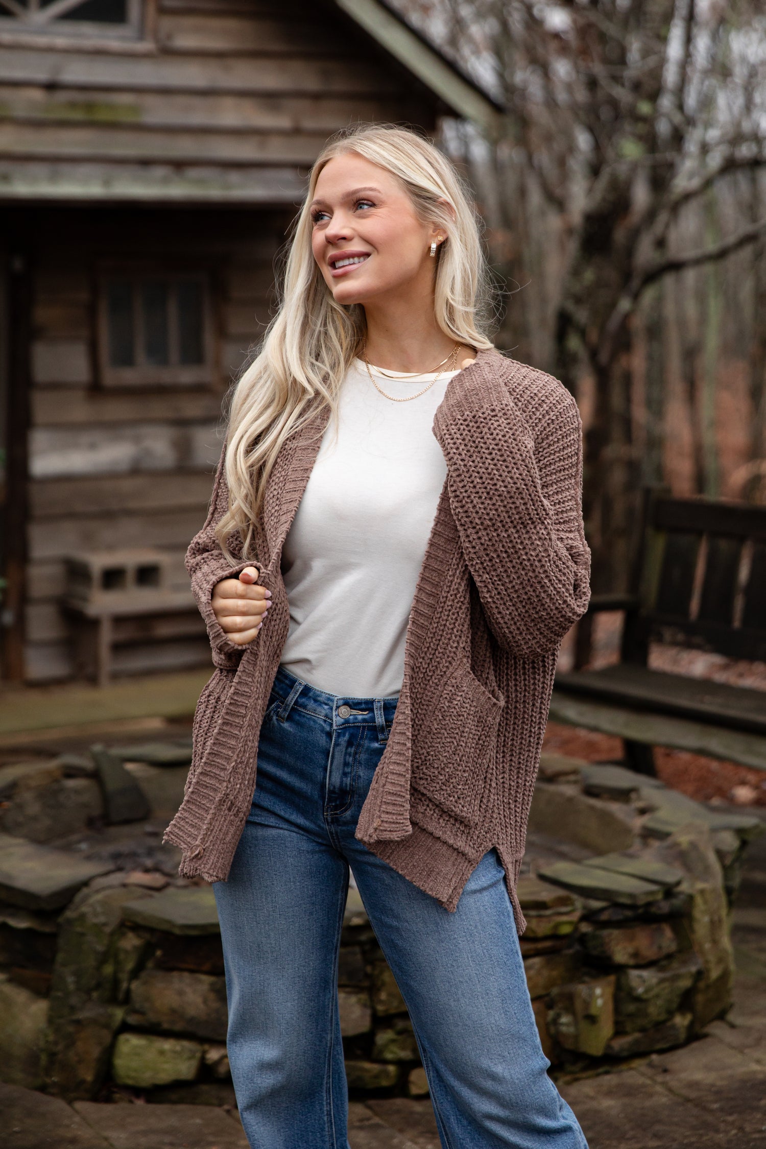 Woman wearing a brown cardigan, white shirt, and blue jeans standing outdoors with a wooden cabin in the background.