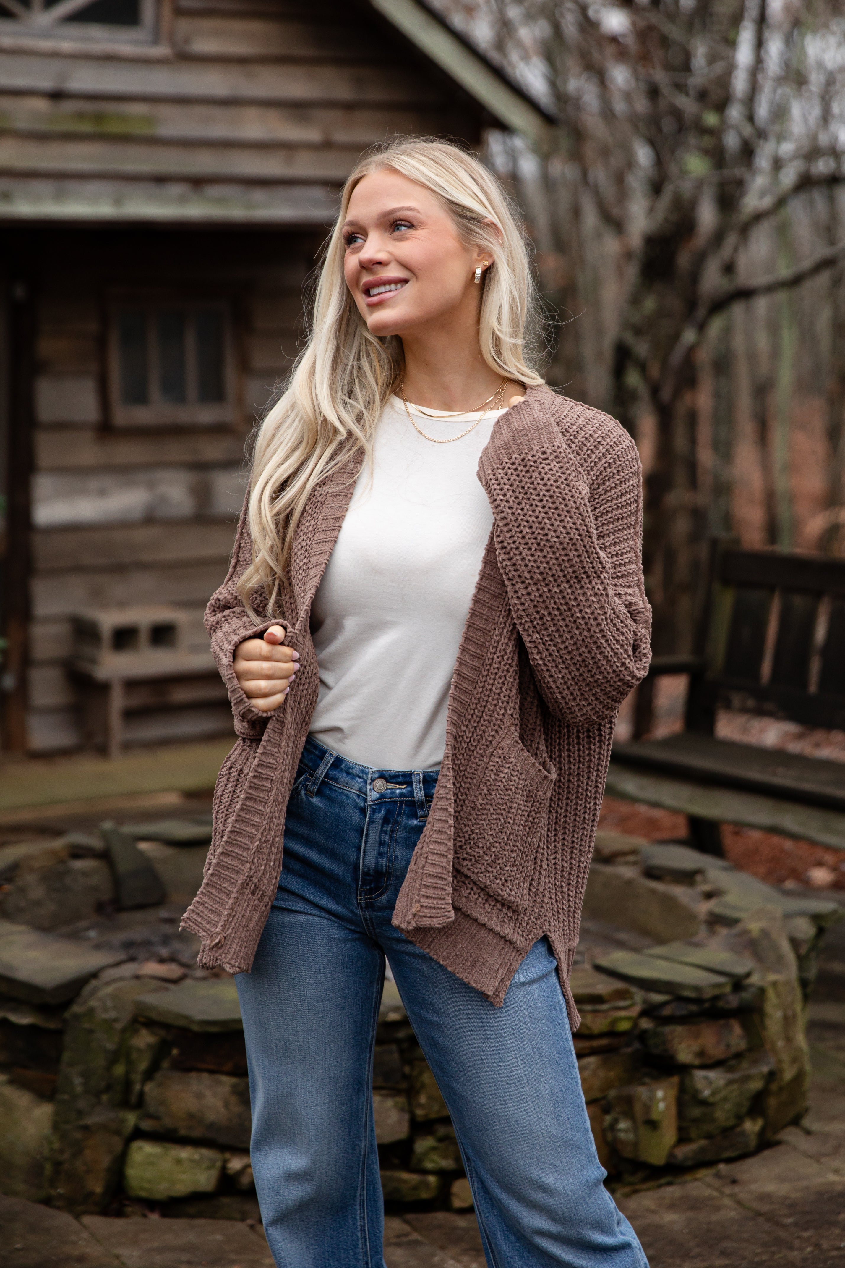 Woman wearing a brown cardigan, white shirt, and blue jeans standing outdoors with a wooden cabin in the background.