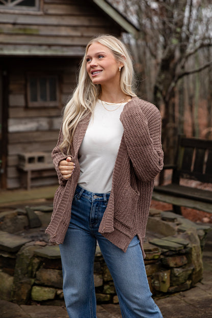 Woman wearing a brown cardigan, white shirt, and blue jeans standing outdoors with a wooden cabin in the background.