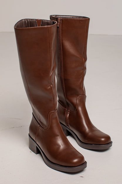 A pair of knee-high brown leather boots with low block heels, shown on a light gray studio background.