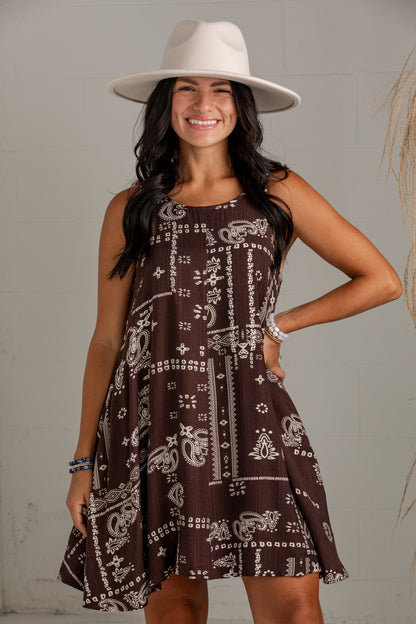 A woman wearing a sleeveless brown dress with a white paisley and geometric pattern, a wide-brimmed cream hat, and bracelets, posing against a light gray brick wall.