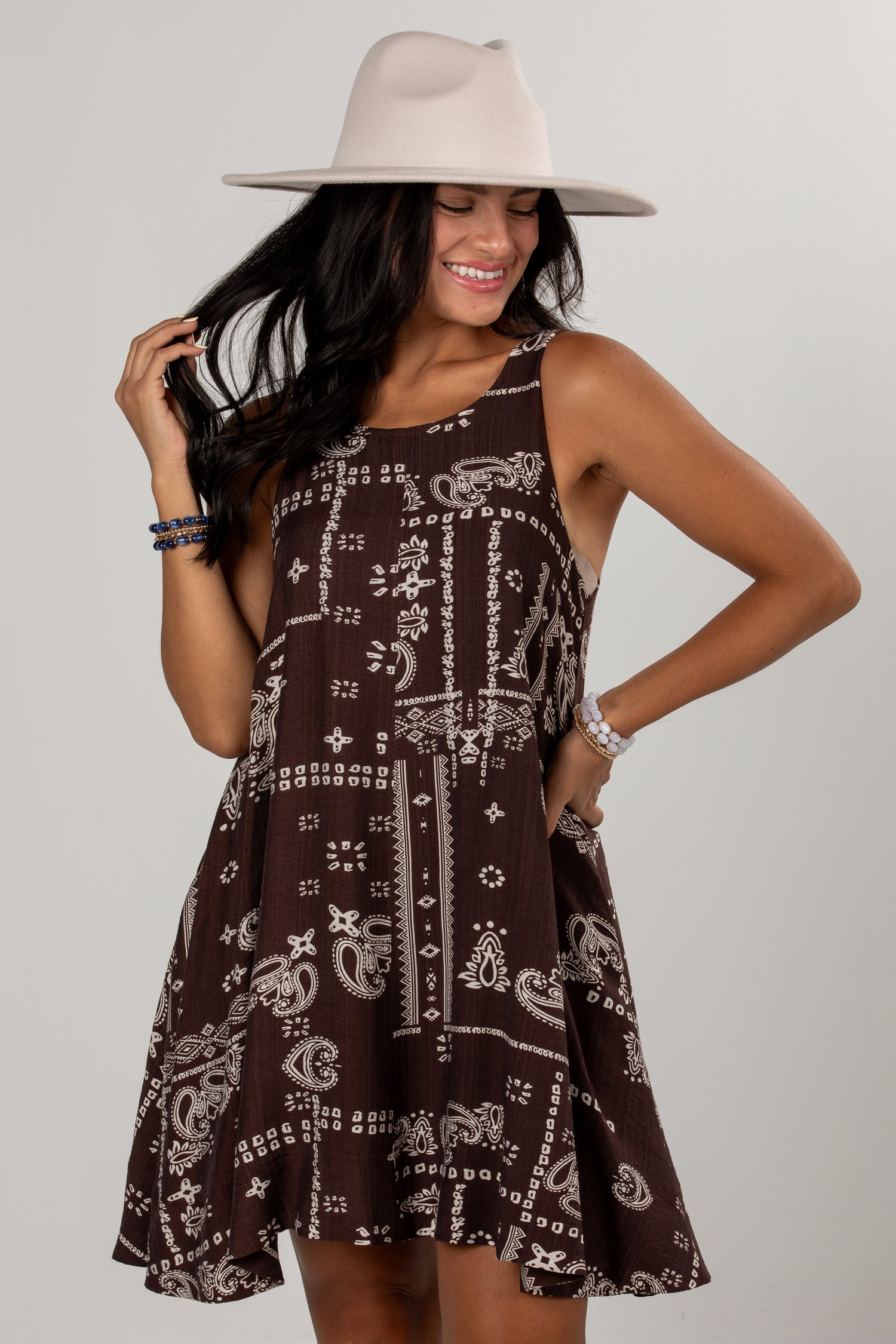 A woman wearing a sleeveless brown dress with a white paisley and bandana-style print, a wide-brim beige hat, and bracelets, posing against a light gray studio background.
