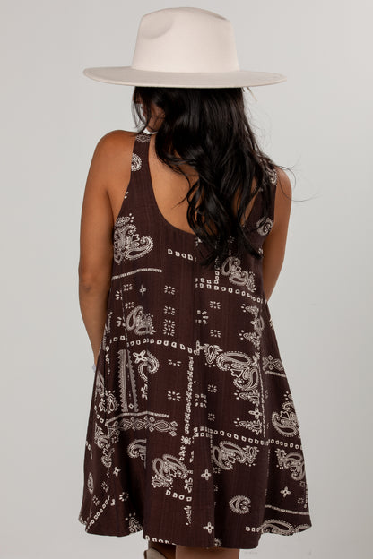 A woman viewed from behind wearing a sleeveless brown dress with a white paisley bandana-style print and a wide-brim cream hat, against a light gray studio background.