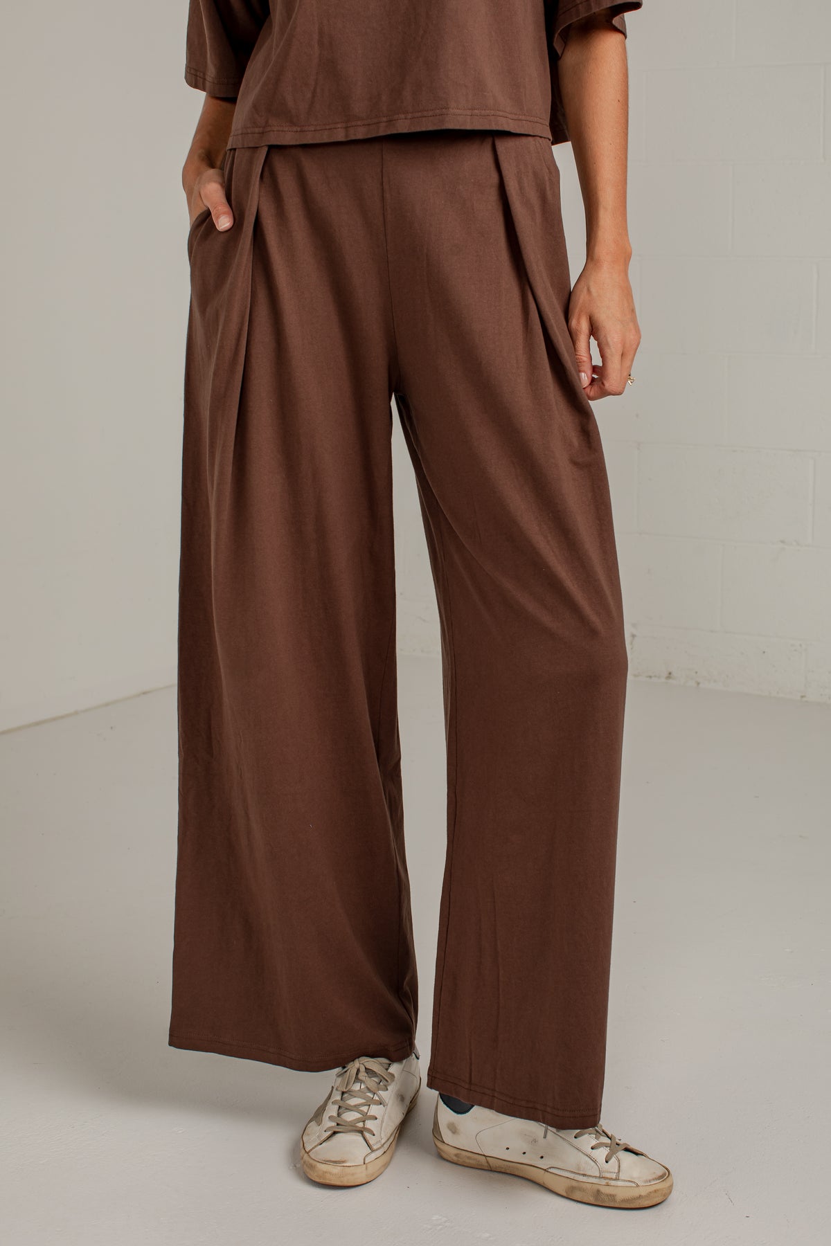 A person wearing loose, wide-leg brown pants with a matching brown top and white sneakers, standing against a plain light gray studio wall.