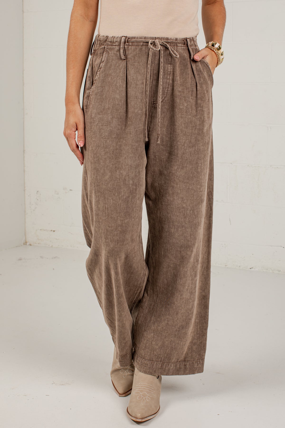 A person wearing high-waisted, wide-leg taupe corduroy pants with a drawstring waist, paired with beige ankle boots in a bright studio setting.