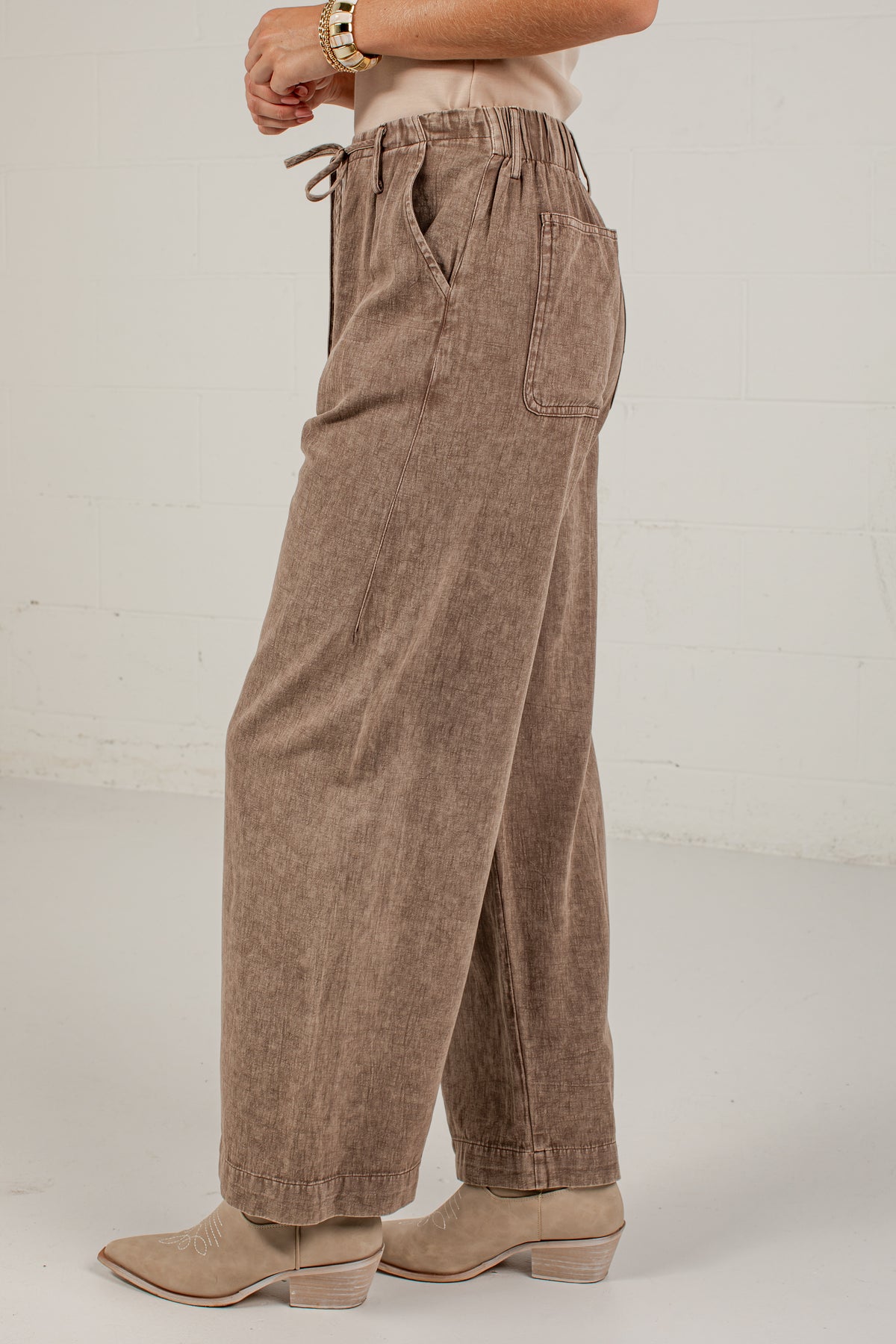 A person shown from the waist down wearing wide-leg brown pants with a drawstring waistband and side pockets, paired with beige ankle boots against a light studio wall.