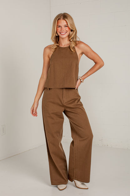 A woman modeling a two-piece brown outfit consisting of a sleeveless halter crop top and matching wide-leg pants, standing against a white studio wall.