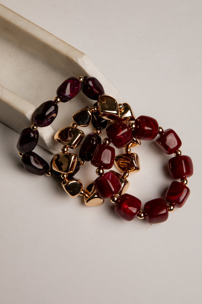 Three beaded stretch bracelets with dark red, burgundy, and black faceted beads separated by gold-toned spacers, displayed on a light beige surface.