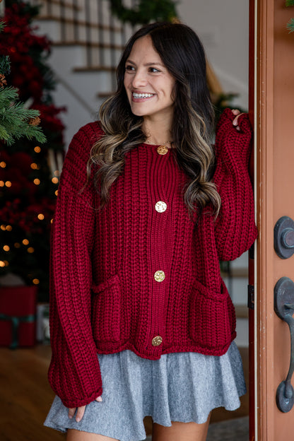 Woman wearing a red knitted cardigan with gold buttons indoors, Christmas decorations in the background.
