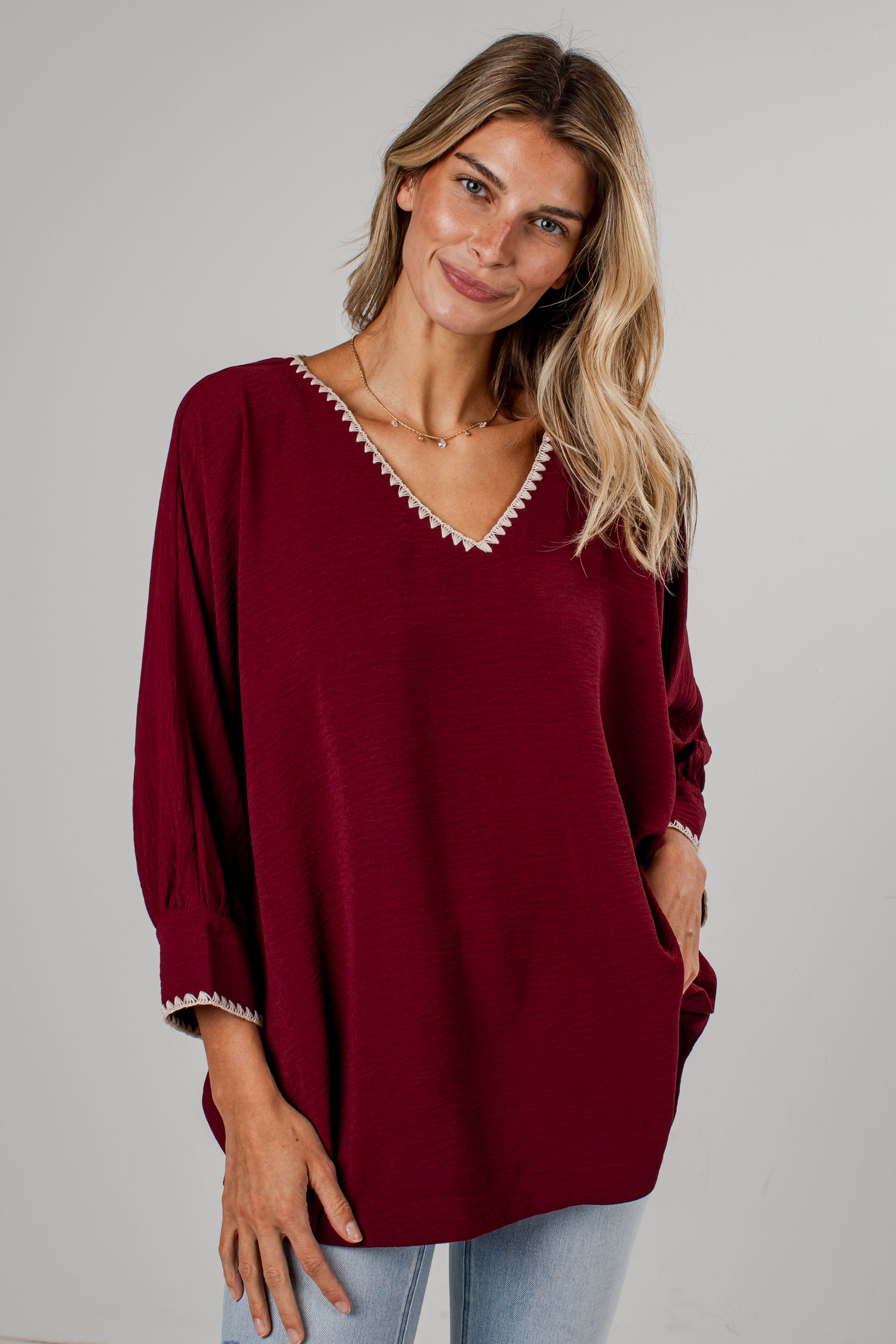 A woman wearing a loose-fitting burgundy blouse with white embroidered trim along the V-neck and cuffs, paired with light-wash blue jeans, standing against a light gray wall.