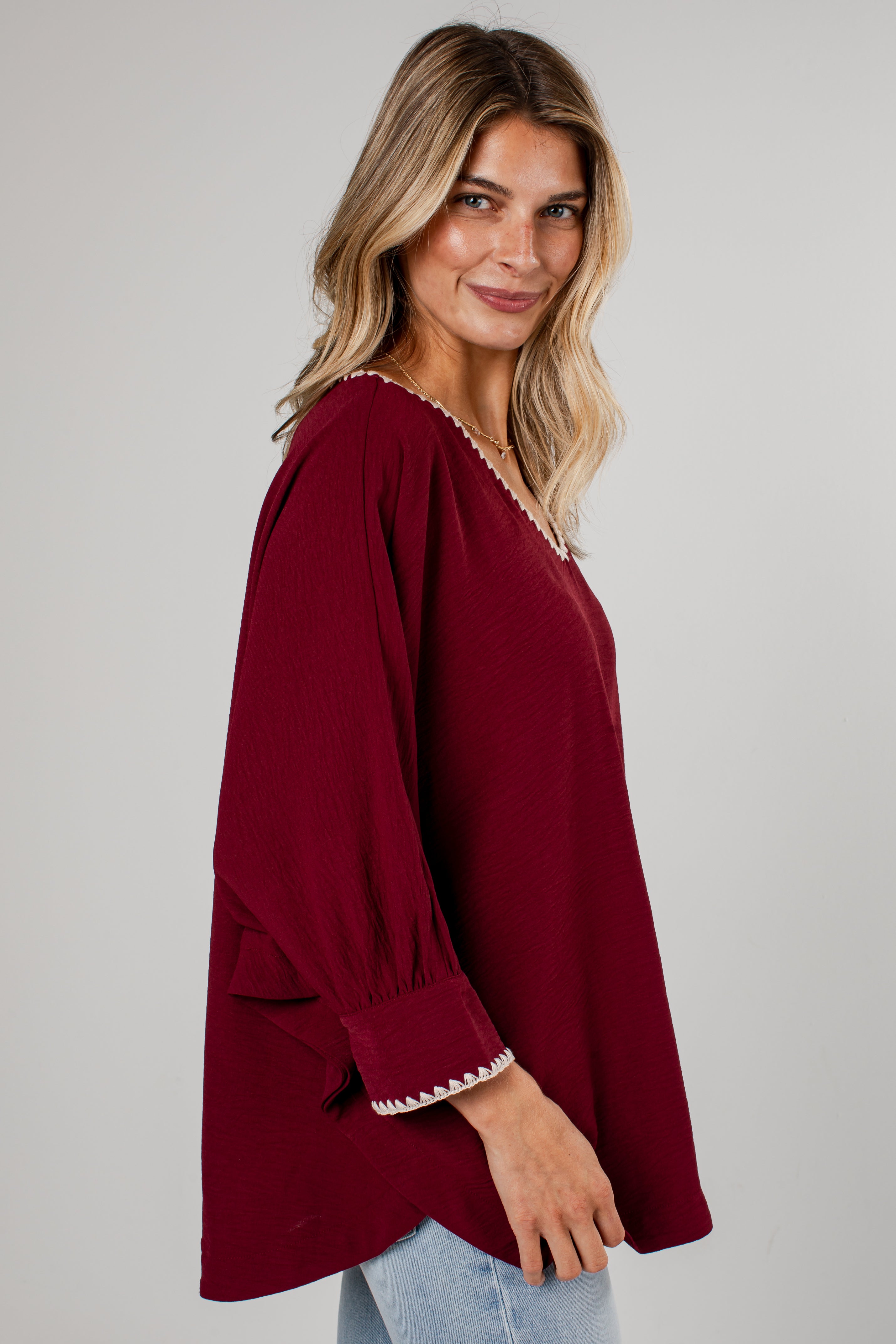 A woman wearing a burgundy long-sleeve top with white embroidered trim on the neckline and cuffs, paired with light blue jeans, photographed against a neutral gray background.