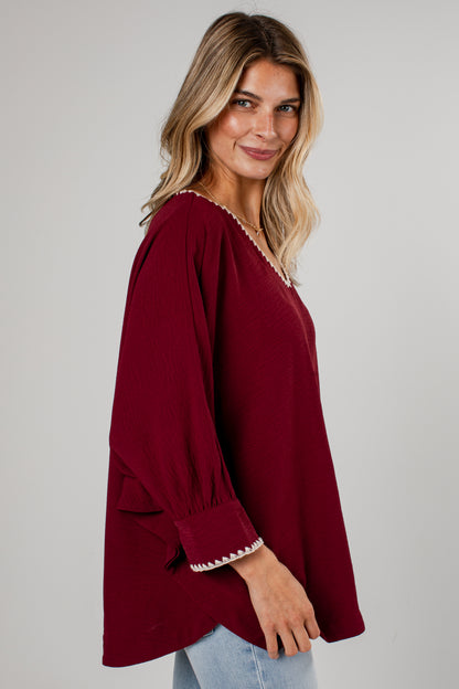 A woman wearing a burgundy long-sleeve top with white embroidered trim on the neckline and cuffs, paired with light blue jeans, photographed against a neutral gray background.