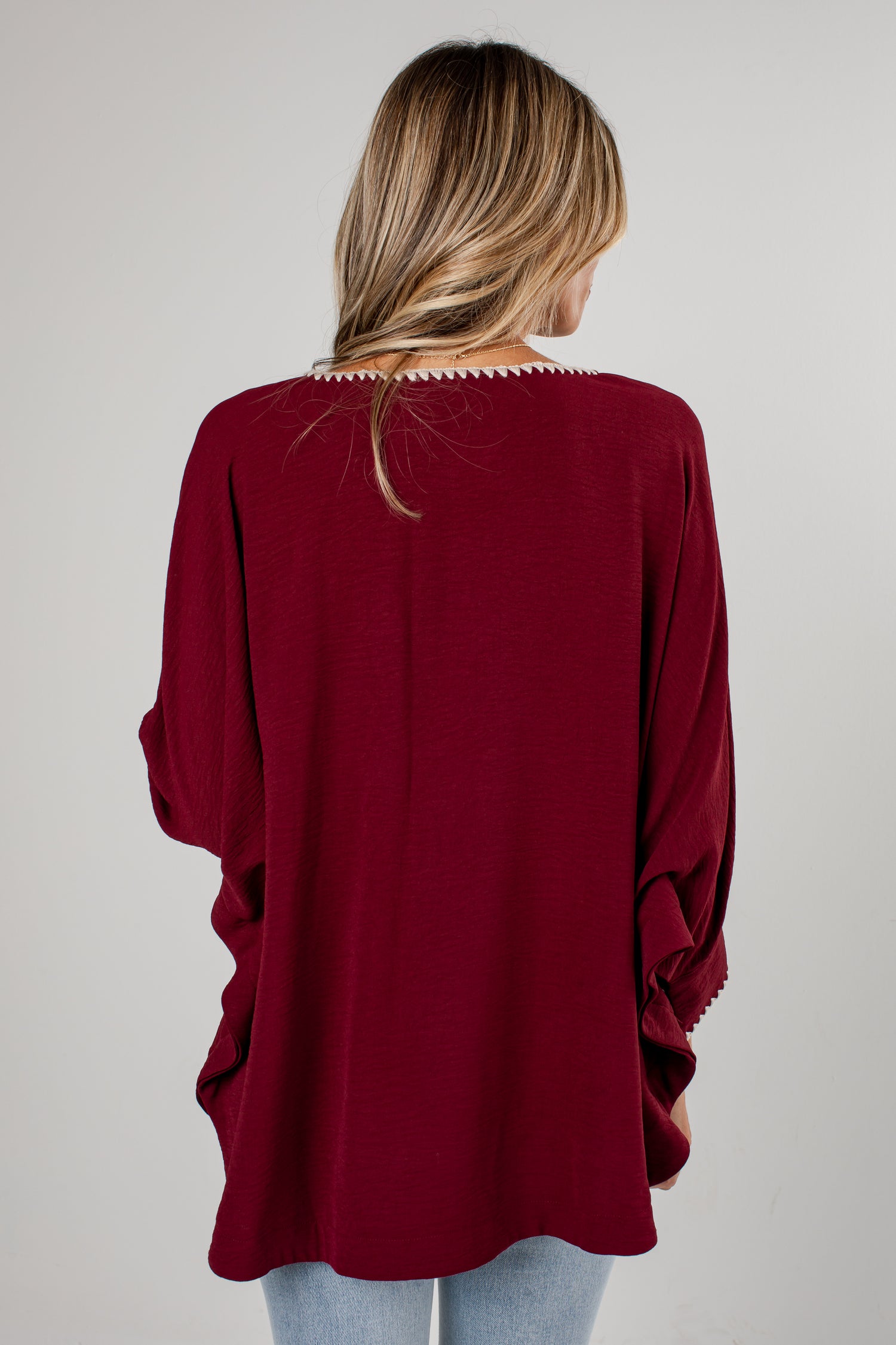 A woman viewed from behind wearing a loose, deep burgundy long-sleeve top with decorative crochet trim around the neckline and sleeve hems, paired with light blue jeans against a plain light gray background.