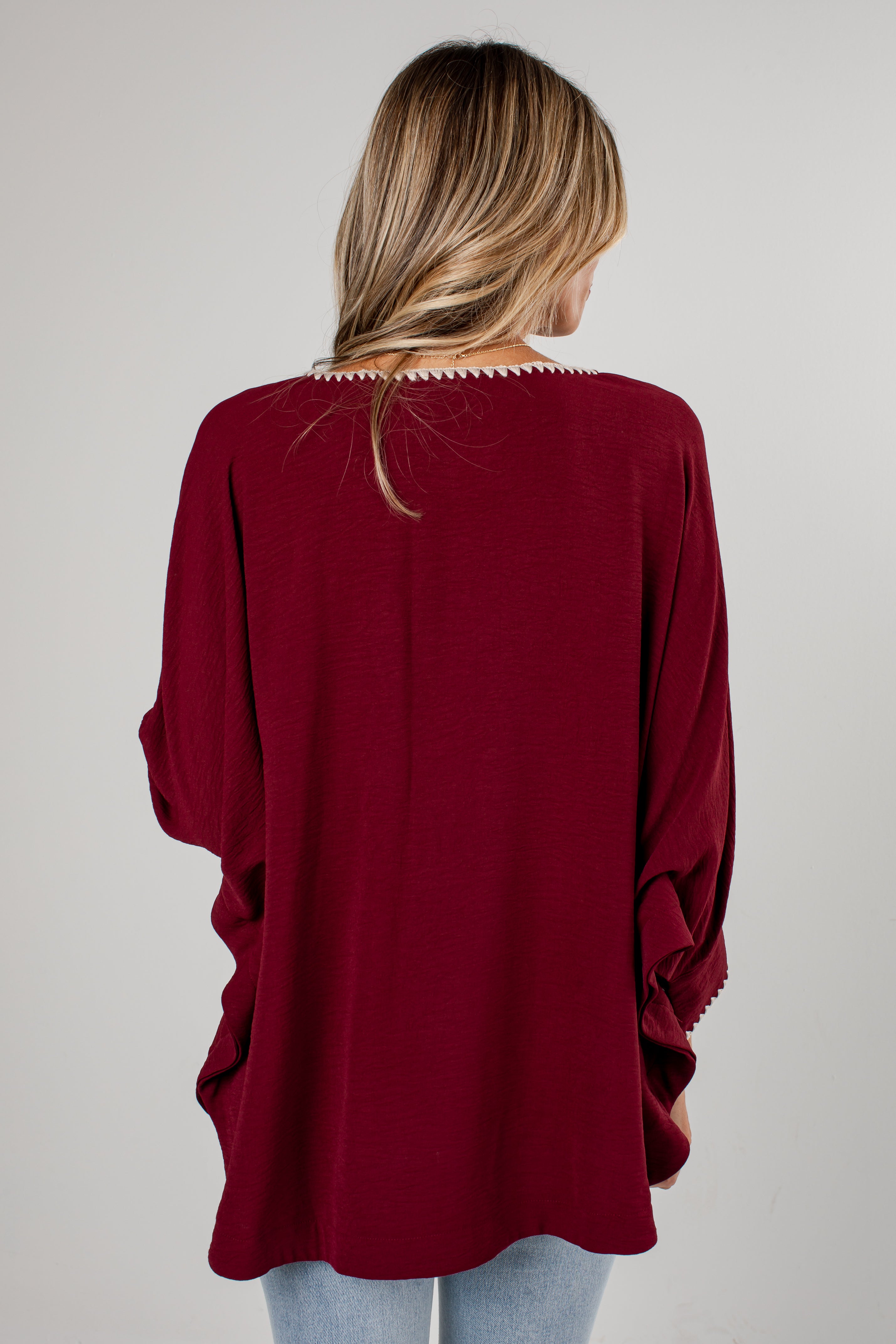 A woman viewed from behind wearing a loose, deep burgundy long-sleeve top with decorative crochet trim around the neckline and sleeve hems, paired with light blue jeans against a plain light gray background.