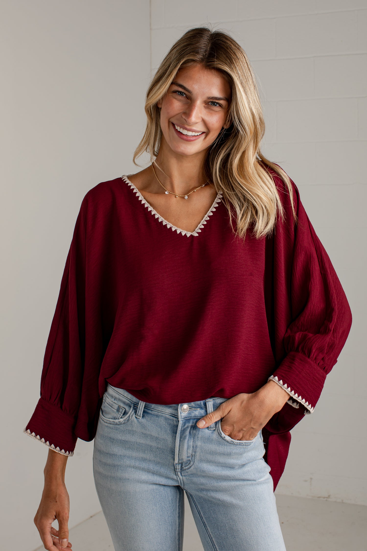 A woman wearing a loose-fitting burgundy blouse with white embroidered trim along the V-neck and cuffs, paired with light-wash blue jeans, standing against a light gray wall.