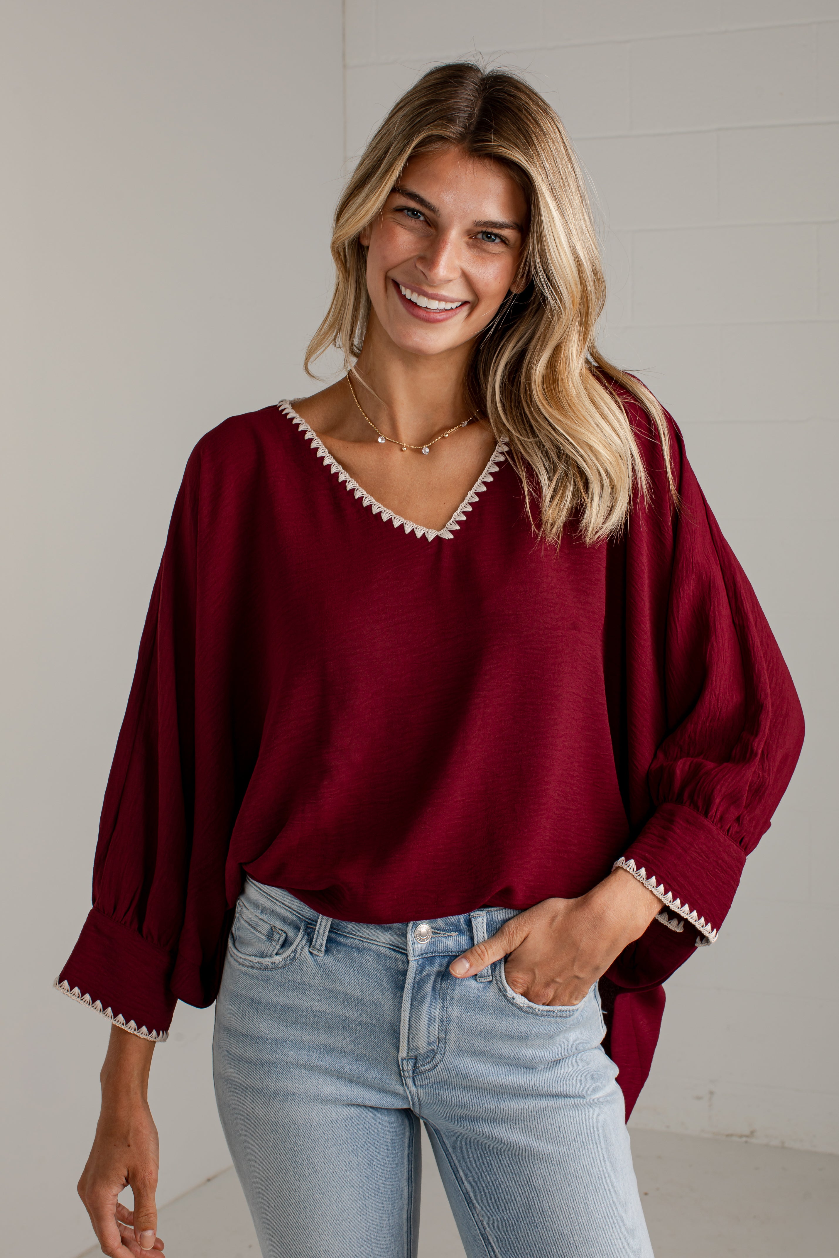 A woman wearing a loose-fitting burgundy blouse with white embroidered trim along the V-neck and cuffs, paired with light-wash blue jeans, standing against a light gray wall.