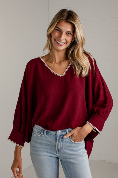 A woman wearing a loose-fitting burgundy blouse with white embroidered trim along the V-neck and cuffs, paired with light-wash blue jeans, standing against a light gray wall.
