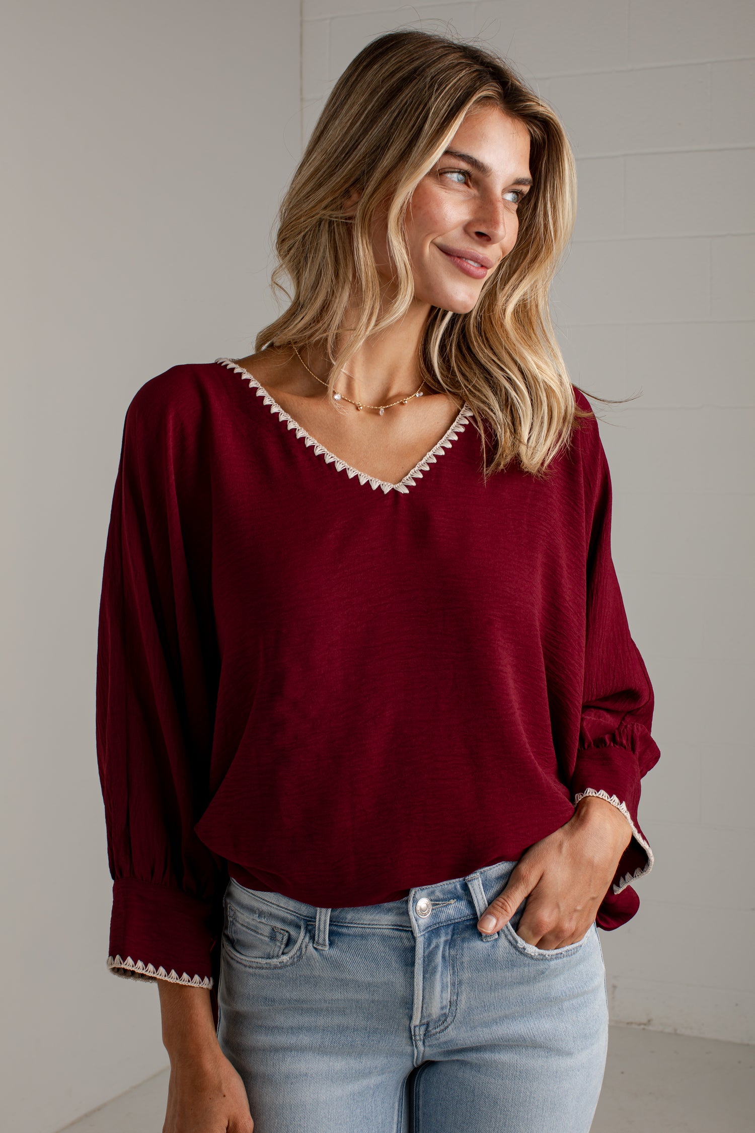 A woman wearing a burgundy long-sleeve blouse with white tassel trim along the V-neck and cuffs, paired with light blue jeans, standing against a light gray wall.