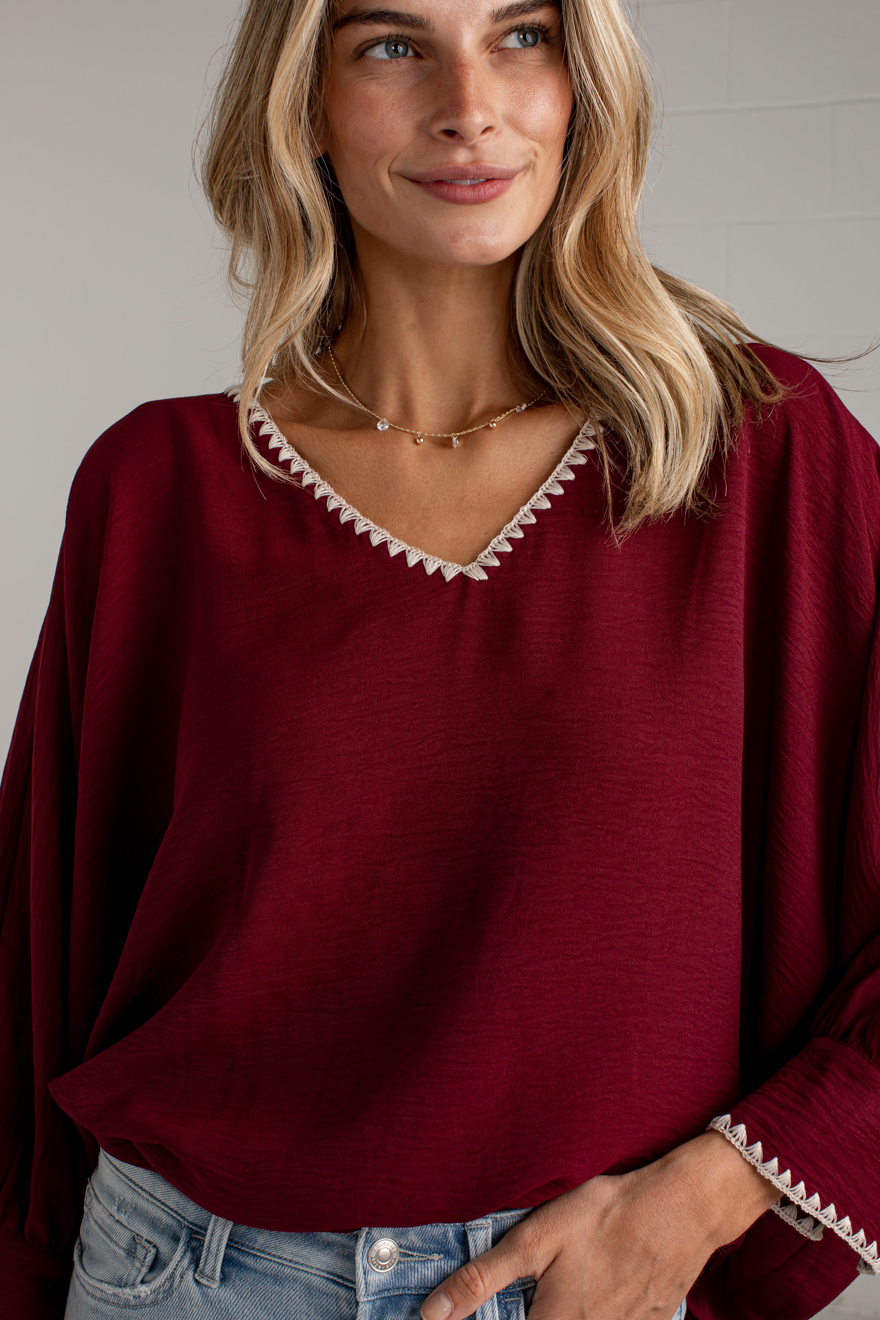 A woman wearing a loose-fitting burgundy blouse with white embroidered trim along the V-neck and cuffs, paired with light-wash blue jeans, standing against a light gray wall.