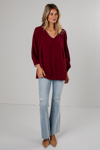 A woman modeling a loose-fitting burgundy long-sleeve top with a decorative white beaded trim along the V-neckline and cuffs, paired with light-wash distressed flare jeans and tan pointed-toe flats, against a plain light gray studio background.