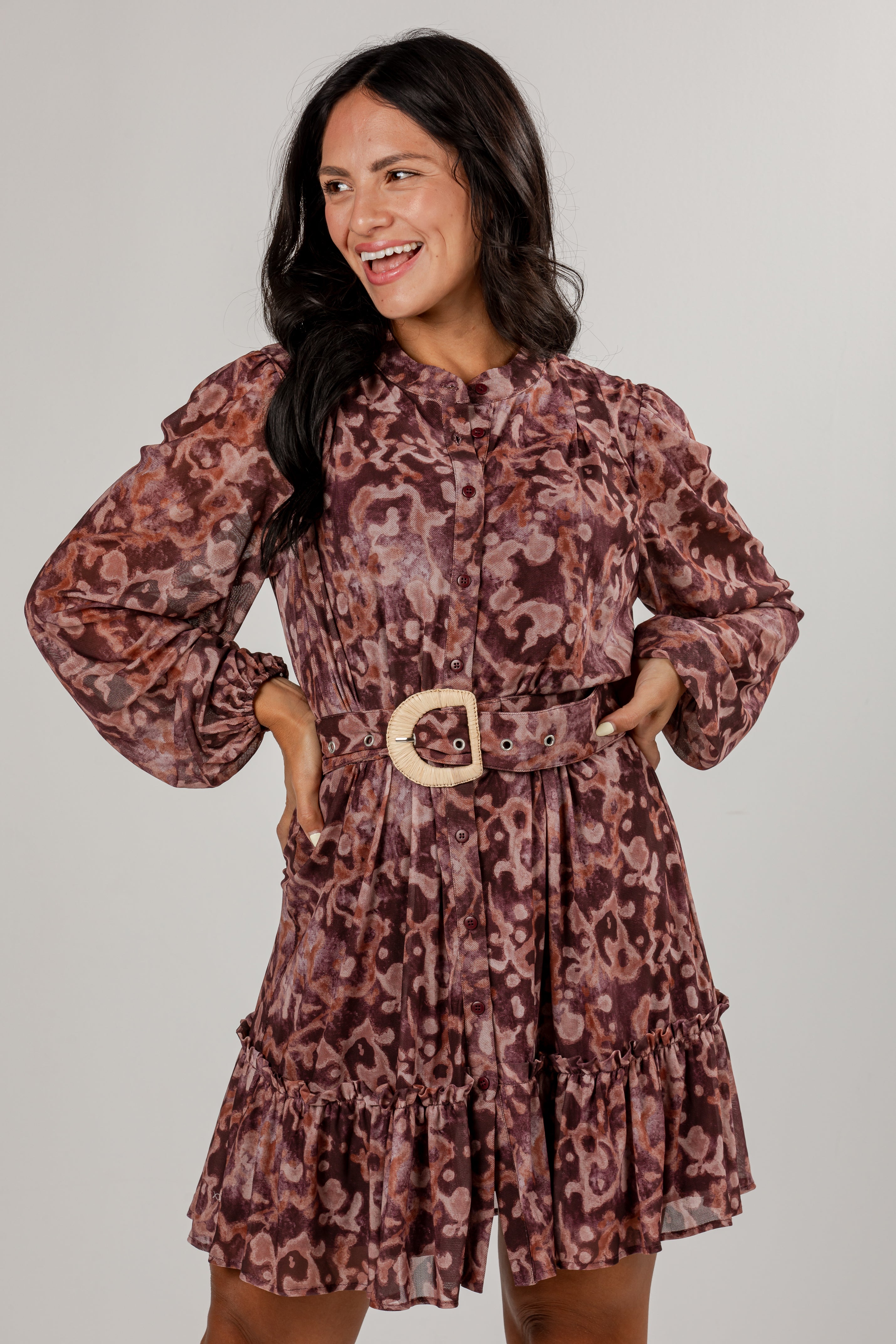 A woman modeling a long-sleeve mini dress with a brown and mauve leopard print, featuring a matching belt with a light-colored wooden buckle, set against a plain light gray background.