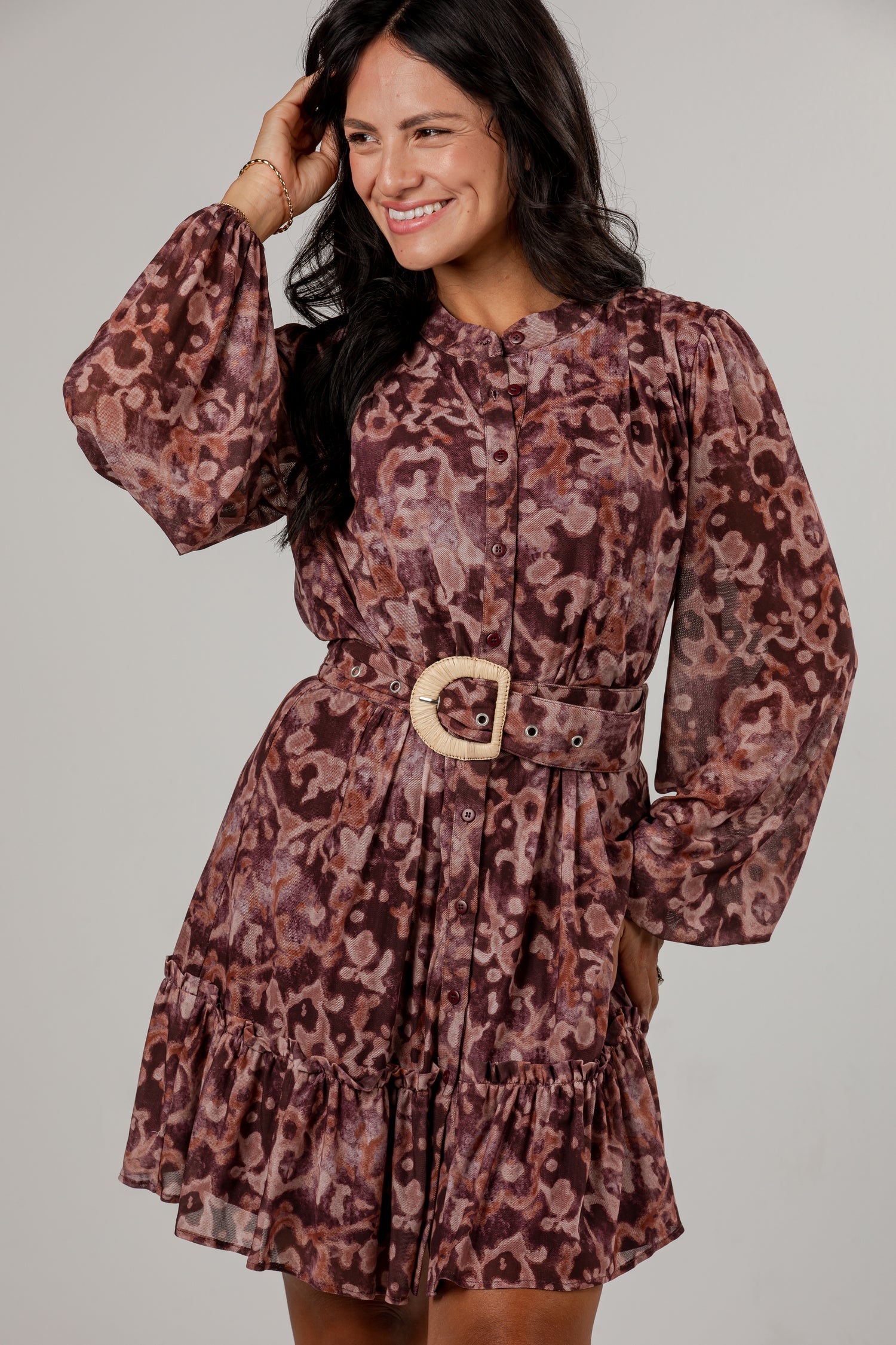 A woman modeling a long-sleeve mini dress with a brown and burgundy abstract pattern, featuring a matching waist belt with a light wood-look buckle, set against a plain light gray studio background.