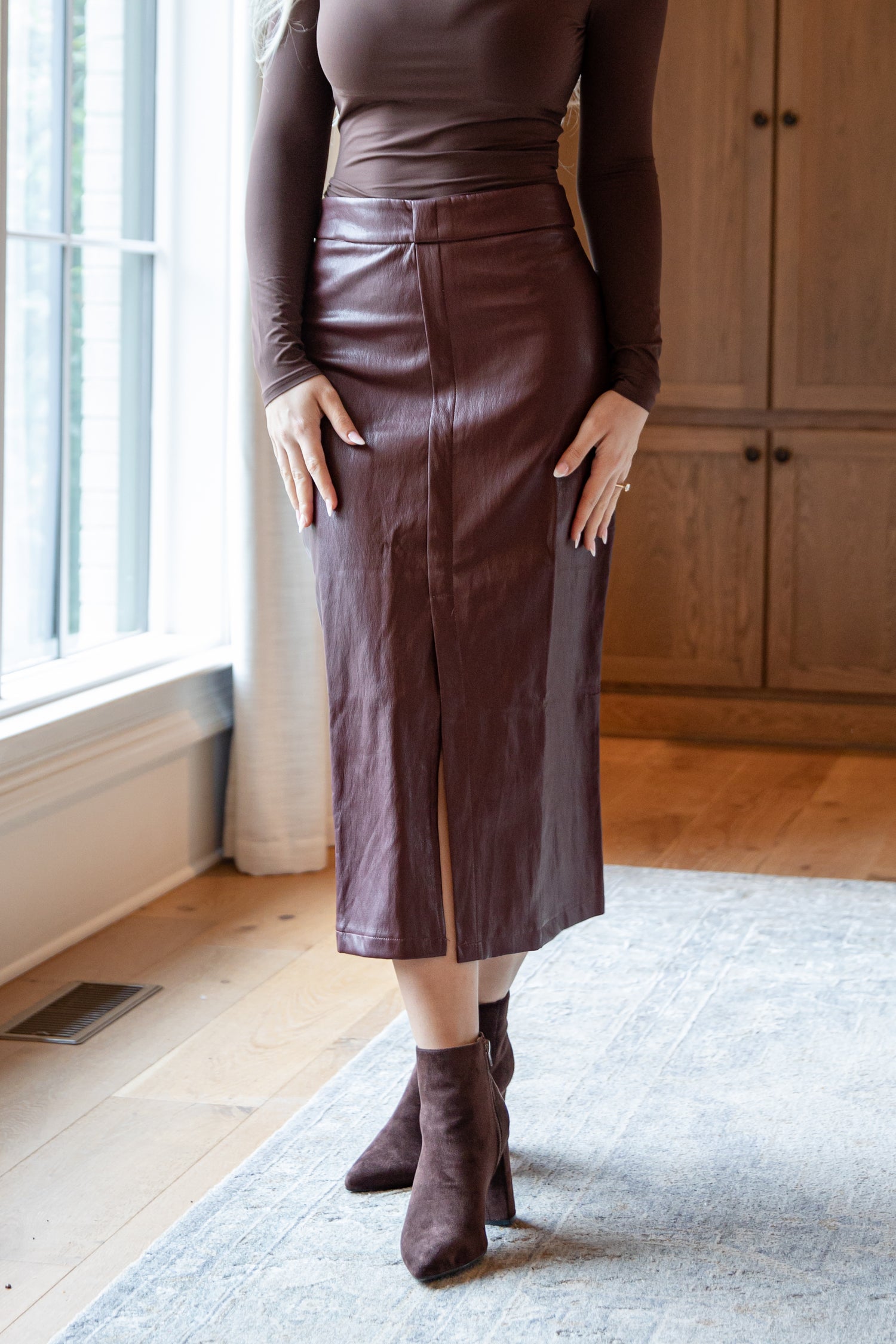 Person wearing a brown leather skirt and matching top in a room with wooden flooring and a window.