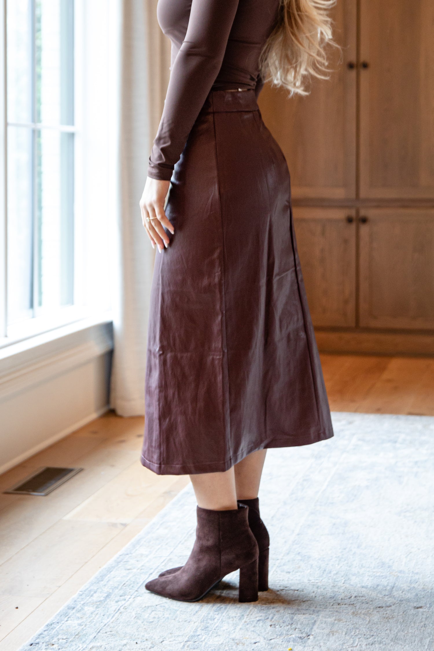 Person wearing a brown leather skirt and matching boots in a room with wooden flooring and a window.