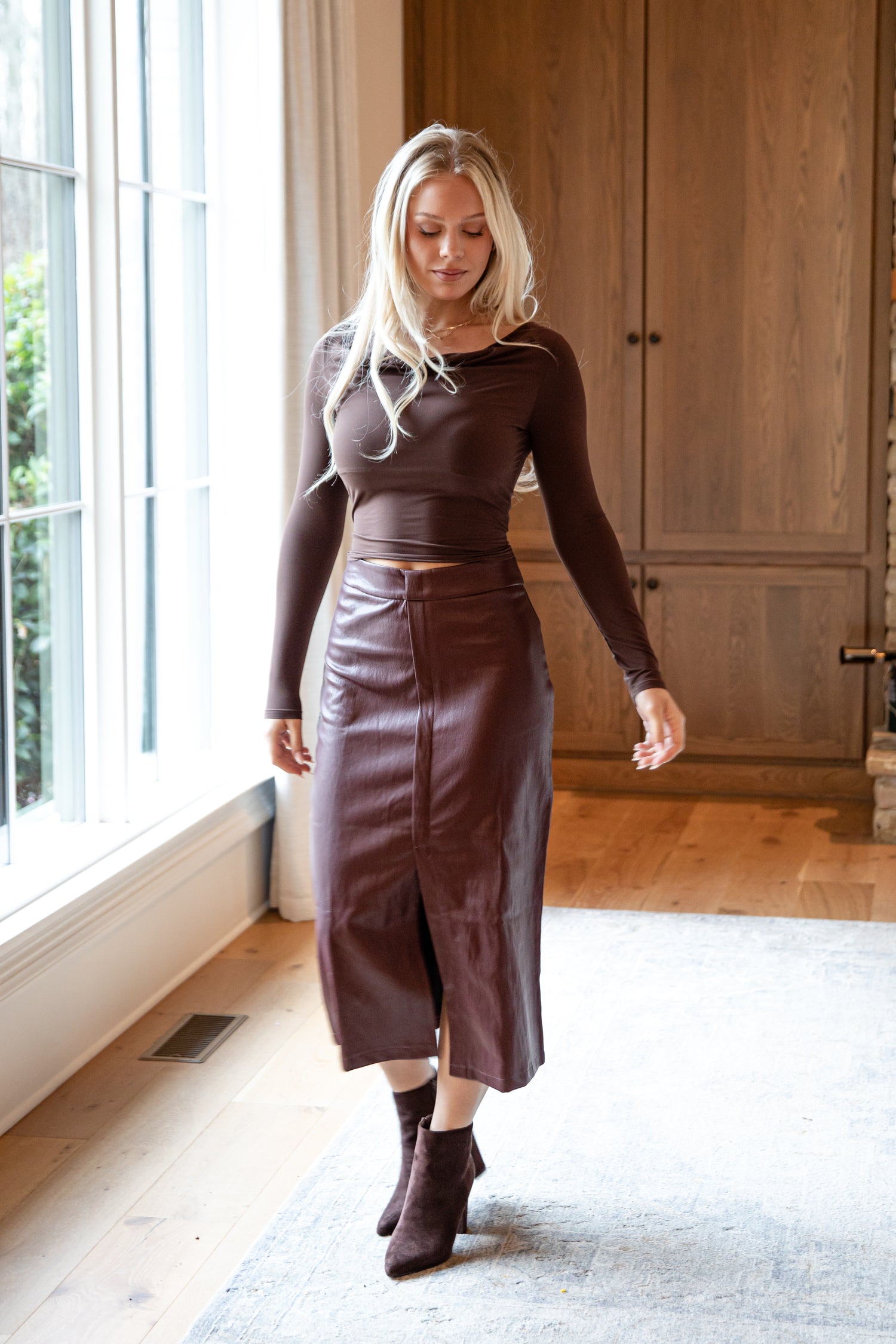 Woman wearing a brown top and leather skirt standing in a room with wooden flooring and large windows.